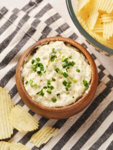 A wooden bowl of creamy white dip garnished with chopped chives, next to crinkle-cut potato chips on a striped cloth.