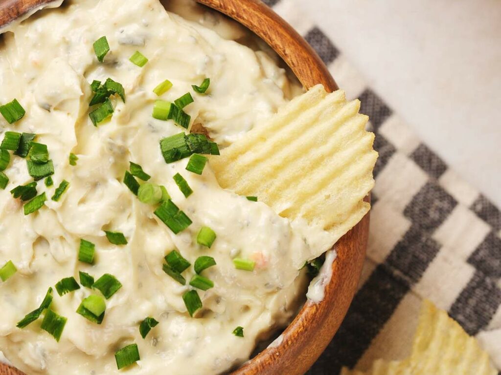 Bowl of creamy dip topped with chopped green onions, accompanied by a ridged potato chip.