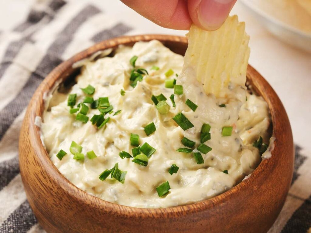 A hand dips a ridged potato chip into a wooden bowl filled with creamy dip topped with chopped green onions.