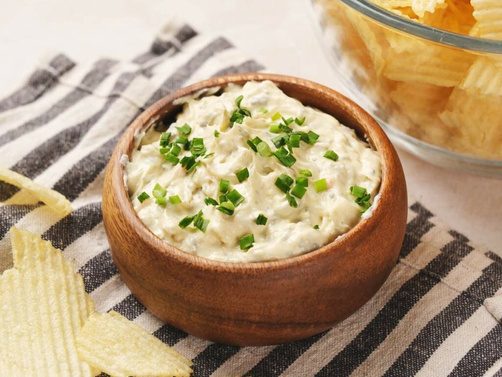 A wooden bowl of creamy chip dip topped with chopped green onions, placed on a striped cloth.