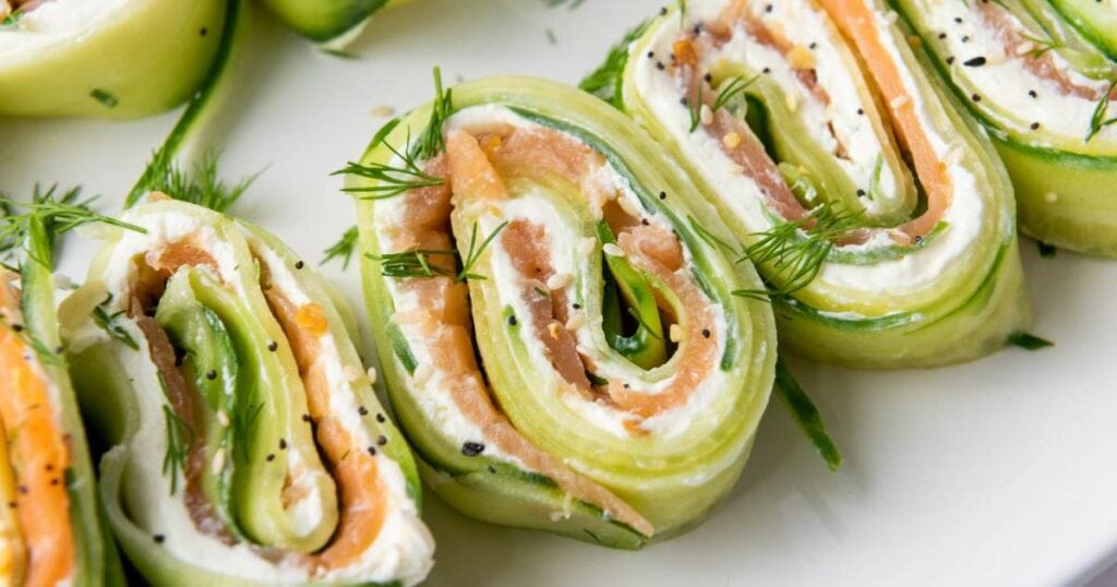 Cucumber appetizers filled with cream cheese and smoked salmon, garnished with fresh dill and black sesame seeds, on a white plate.