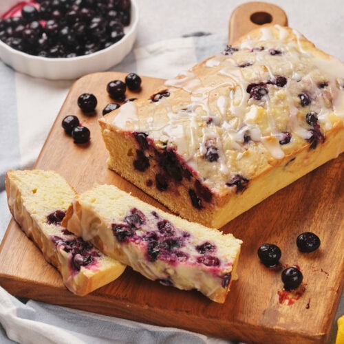 Loaf of blueberry bread with icing on a wooden cutting board, surrounded by fresh blueberries and lemons. Two slices are cut from the loaf.