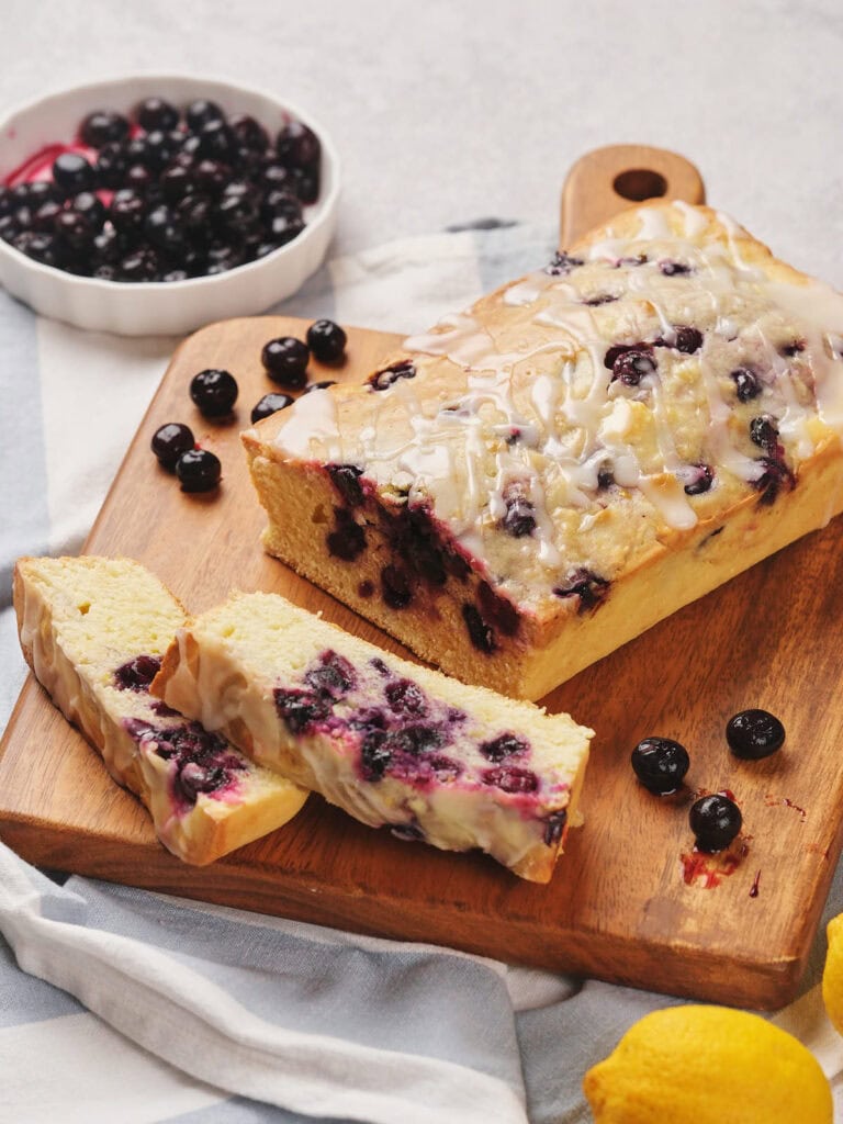 Loaf of blueberry bread with icing on a wooden cutting board, surrounded by fresh blueberries and lemons. Two slices are cut from the loaf.