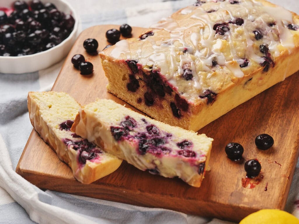 A loaf of blueberry bread with icing sits on a wooden board. Two slices are cut, revealing blueberries inside. A bowl of blueberries is in the background.