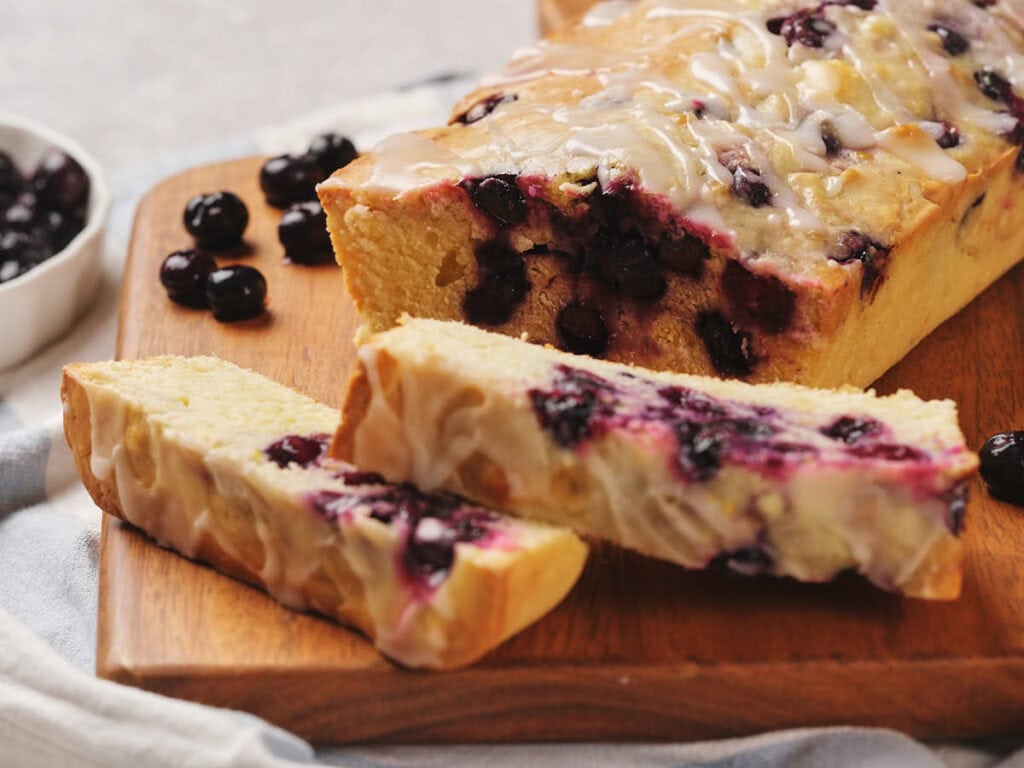 A sliced blueberry loaf cake with a glaze on a wooden board. A small bowl of blueberries is in the background.