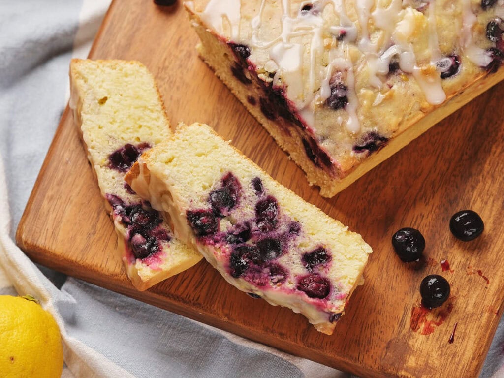 Sliced blueberry lemon bread with icing drizzled on top, arranged on a wooden board. Nearby are whole blueberries and a lemon.