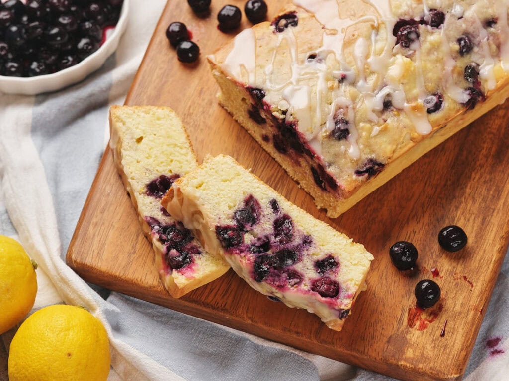 Sliced blueberry lemon loaf with icing on a wooden board, surrounded by fresh blueberries and two lemons.