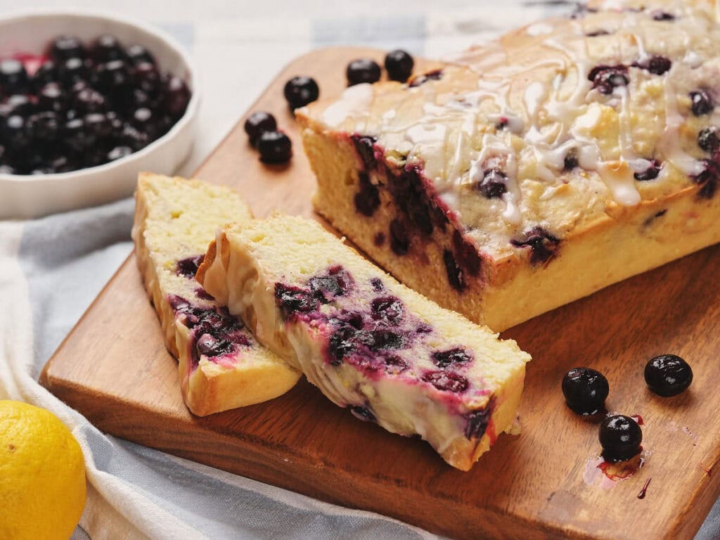 Sliced blueberry loaf cake with glaze on a wooden board, accompanied by a bowl of blueberries and a lemon.