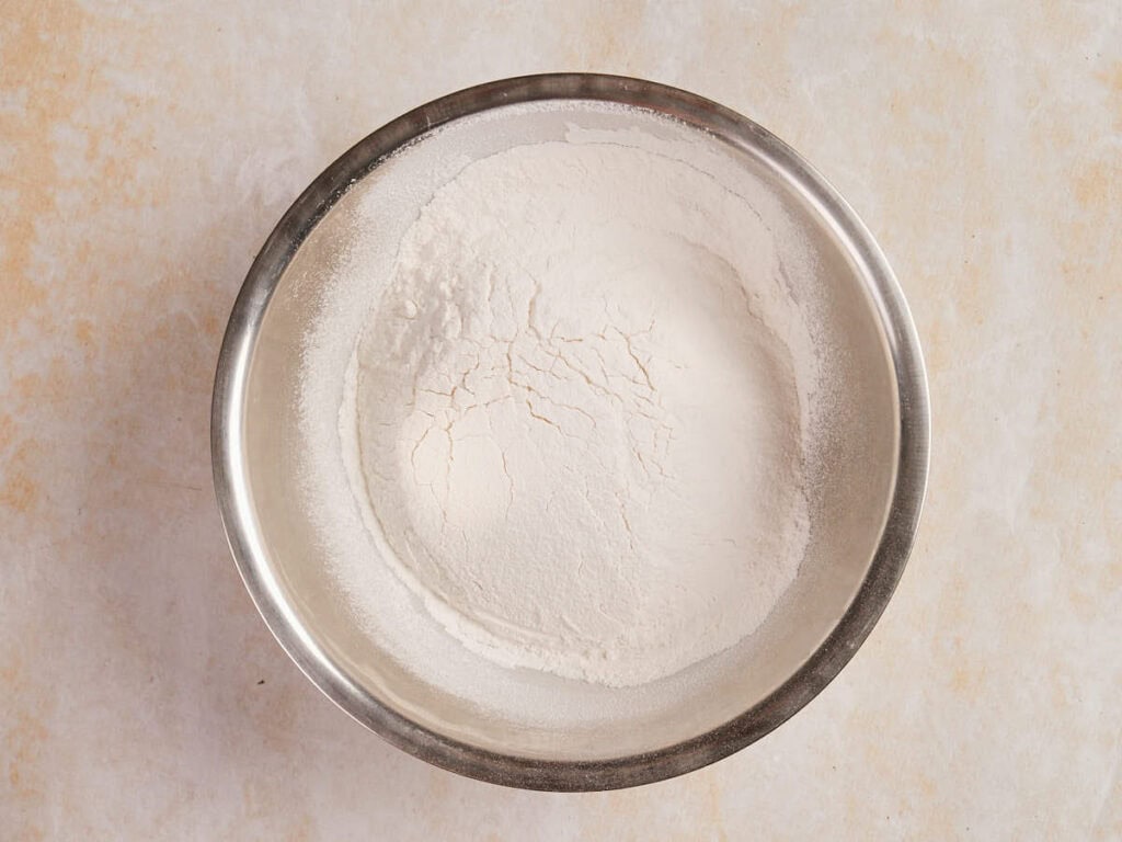 A metal bowl filled with white flour on a light textured surface.