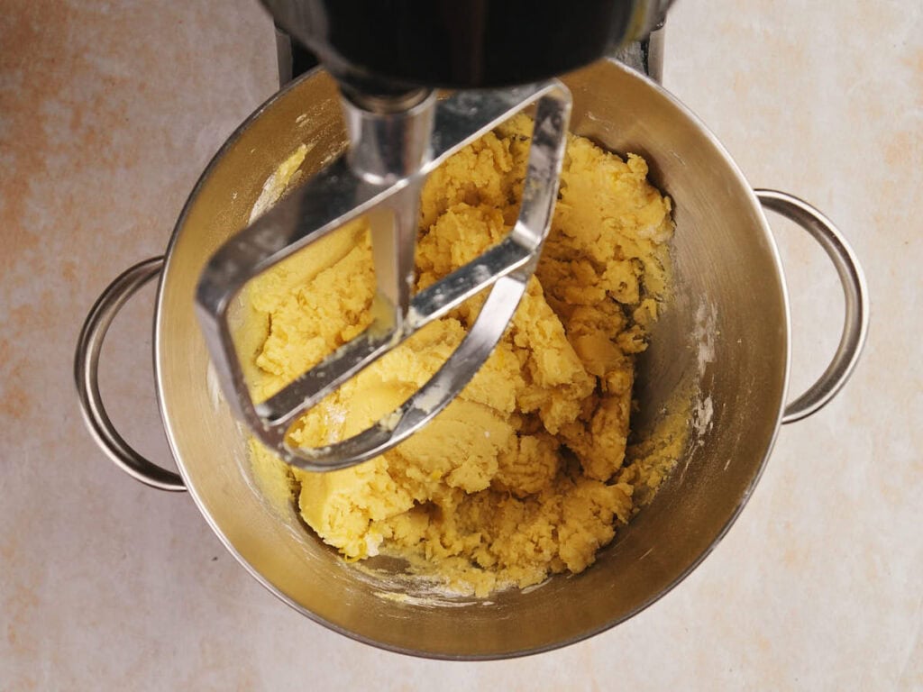 A mixing bowl with yellow dough and a paddle attachment in a stand mixer.