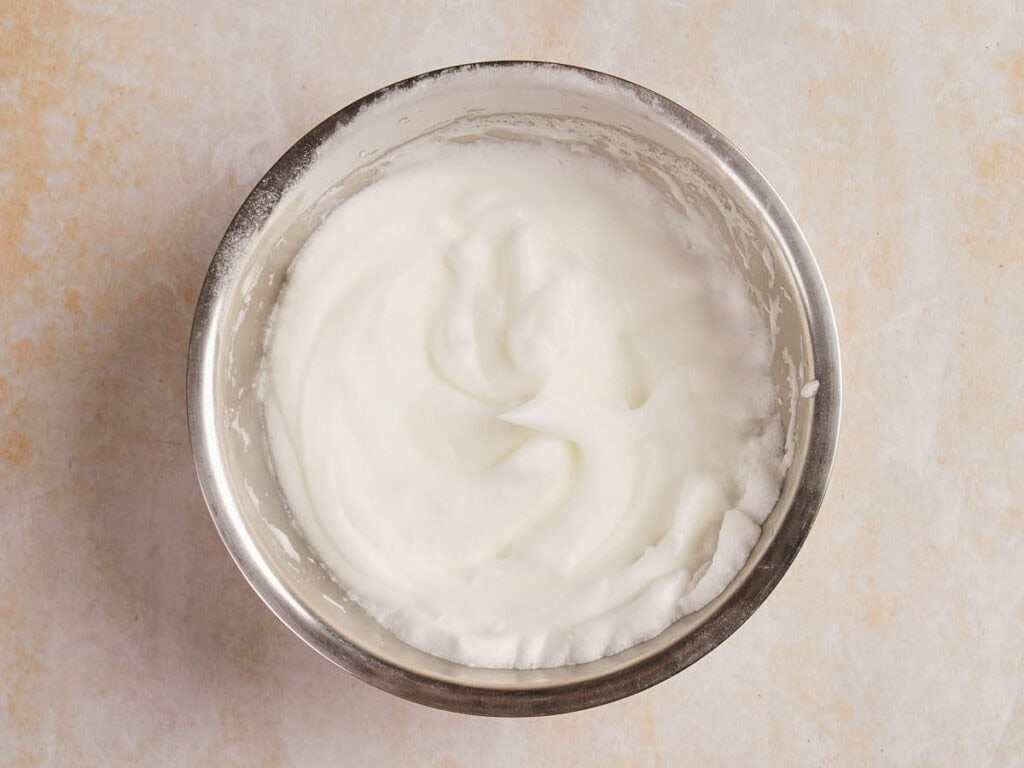 A stainless steel bowl filled with whipped egg whites on a light-colored surface.