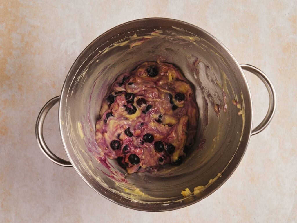 A metal bowl containing a mixture of dough with blueberries is placed on a light-colored surface.