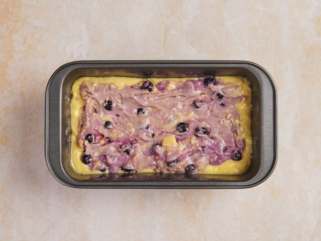 Loaf pan with blueberry swirl batter on a light-colored surface.