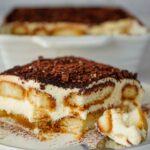 A slice of tiramisu on a plate with a fork beside it, showing layers of cream and coffee-soaked biscuits. A full dish of tiramisu is in the background.