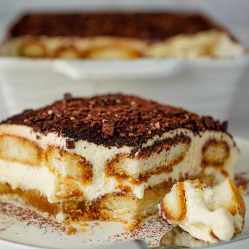A slice of tiramisu on a plate with a fork beside it, showing layers of cream and coffee-soaked biscuits. A full dish of tiramisu is in the background.
