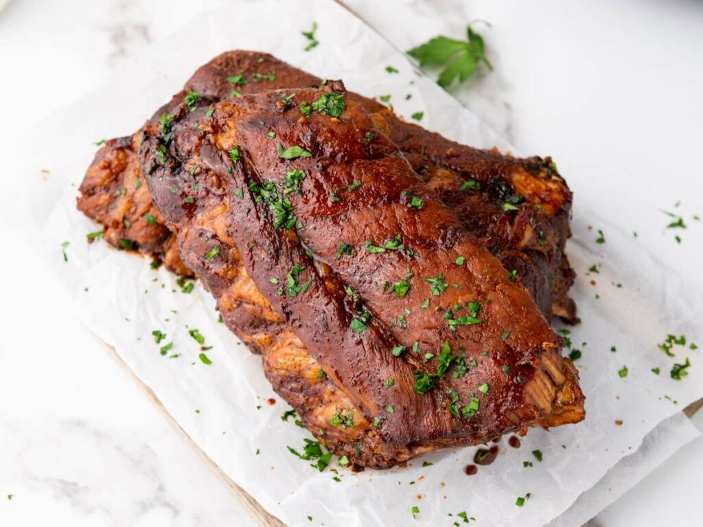 Baby back ribs stacked on parchment paper, garnished with chopped parsley, on a white countertop.