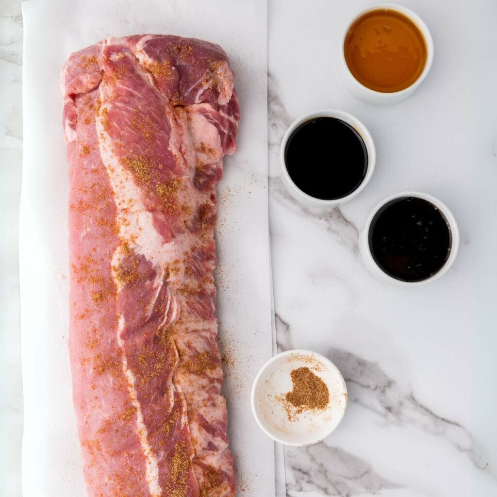 Raw pork tenderloin sprinkled with seasoning on parchment paper, next to four small bowls containing various sauces and spices.
