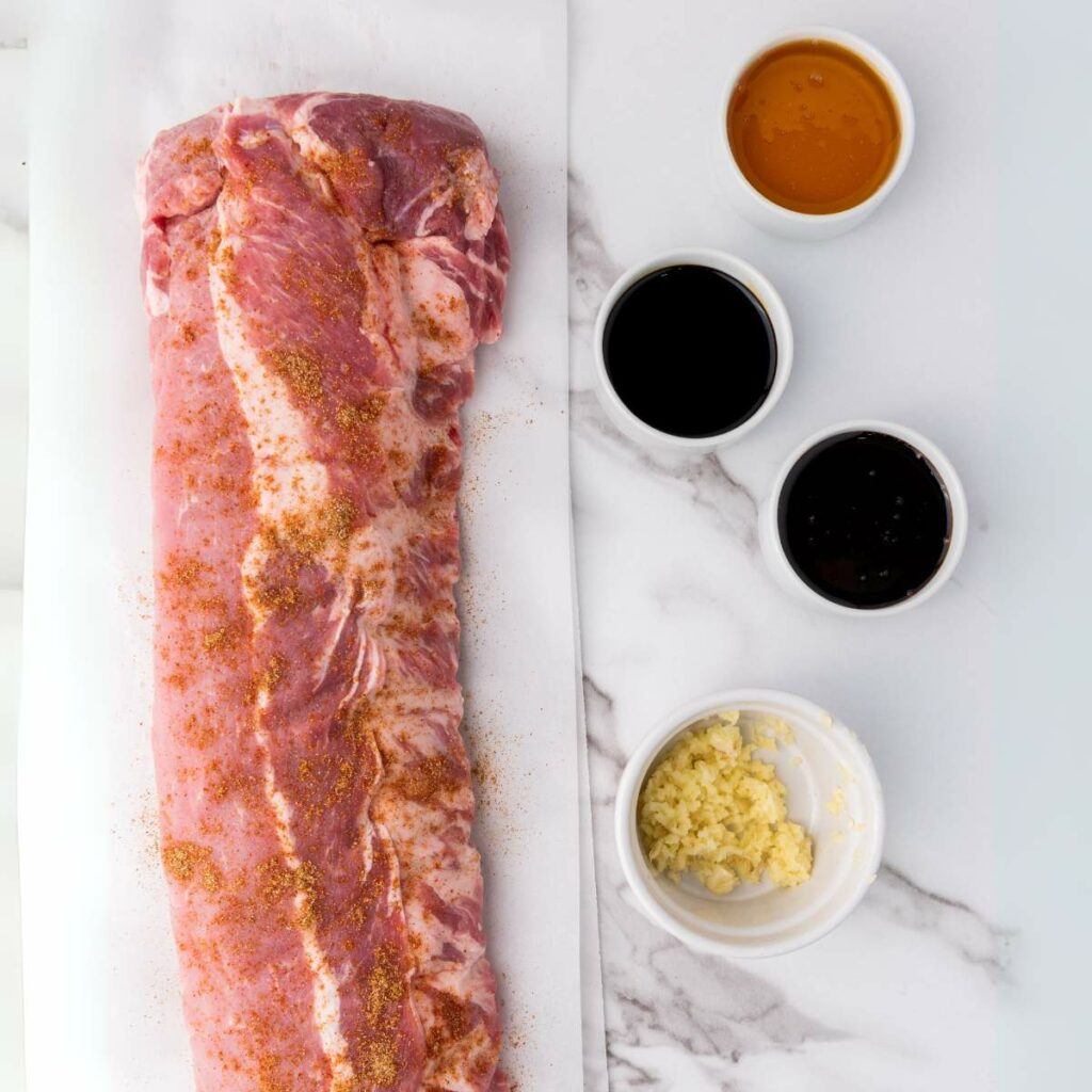 Raw pork ribs seasoned with spices are placed on parchment paper next to four small bowls.
