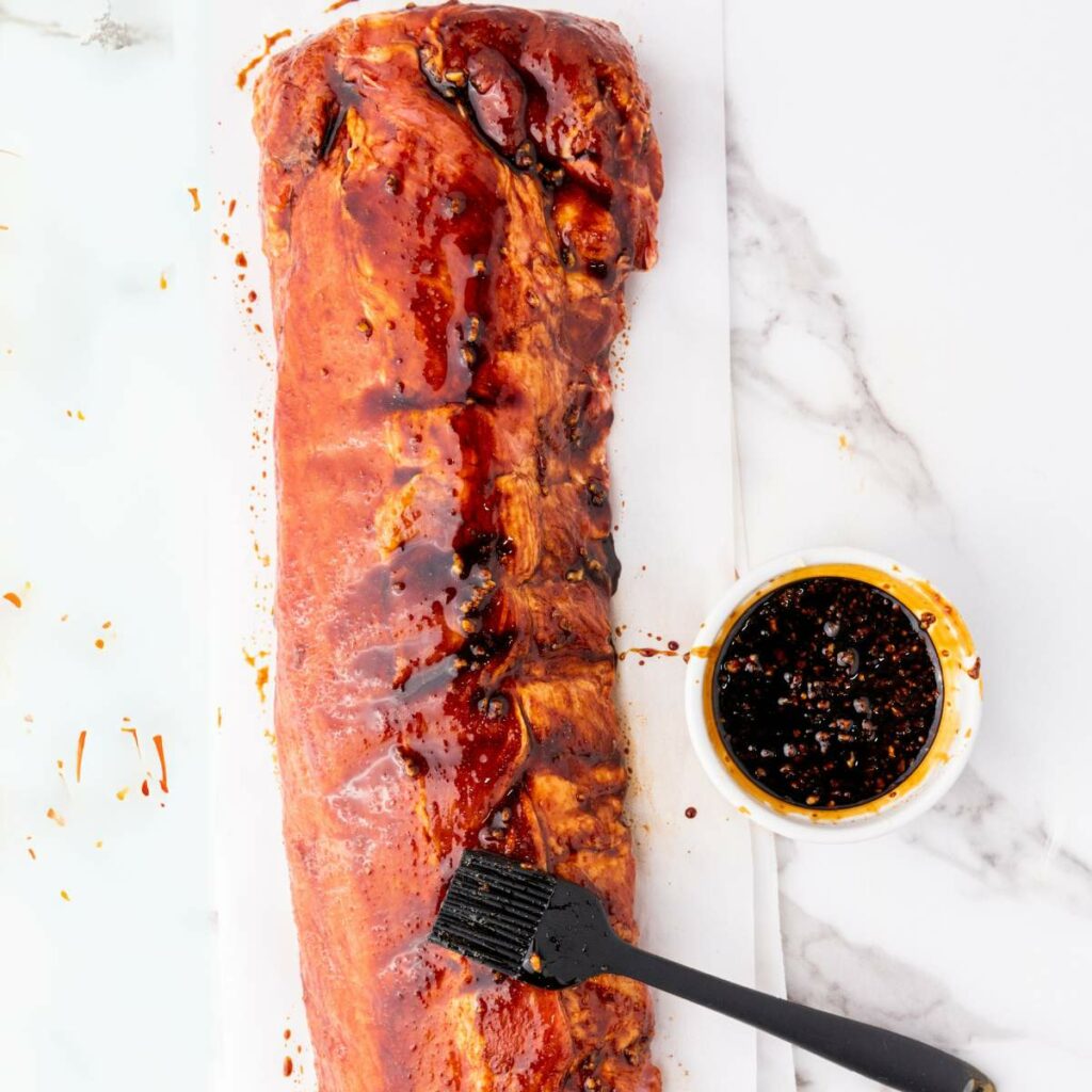 A rack of pork ribs is being brushed with a thick, dark sauce next to a small bowl of sauce on a white marble surface.