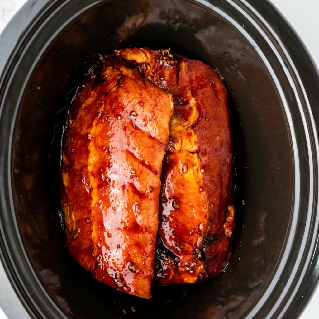 Two pieces of glazed meat are shown cooking in a black slow cooker, coated with a shiny, dark sauce.