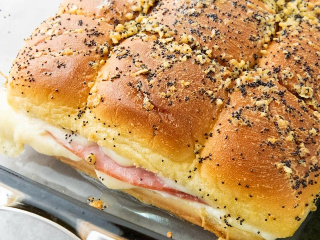 Baked ham and cheese sliders topped with poppy seeds and seasonings in a glass dish.