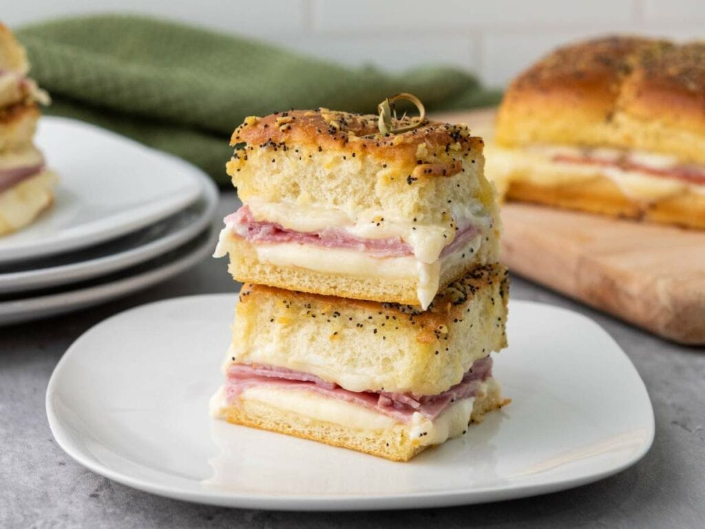 Stacked ham and cheese sliders on a white plate with poppy seeds on top.