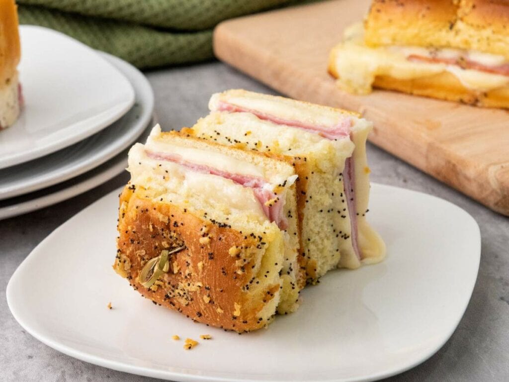 Two ham and cheese sliders on an open bun are topped with a buttery poppy seed dressing on a white plate. A green cloth and more sliders on a cutting board are in the background.