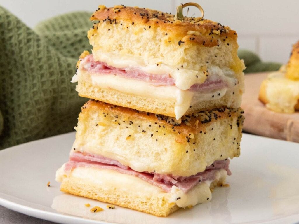 Two stacked ham and cheese sliders with poppy seeds on top, served on a white plate.