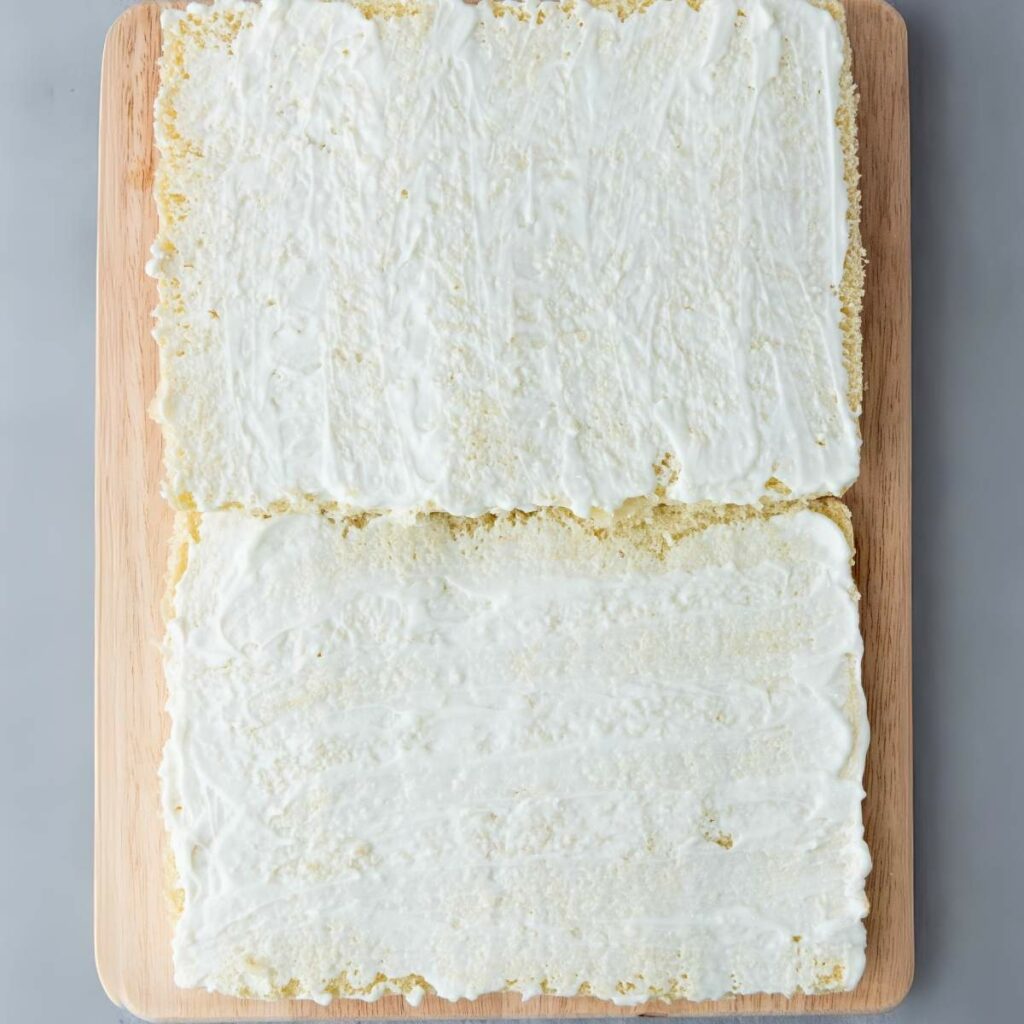 Two rectangular layers of cake covered with white frosting on a wooden cutting board, placed on a gray surface.