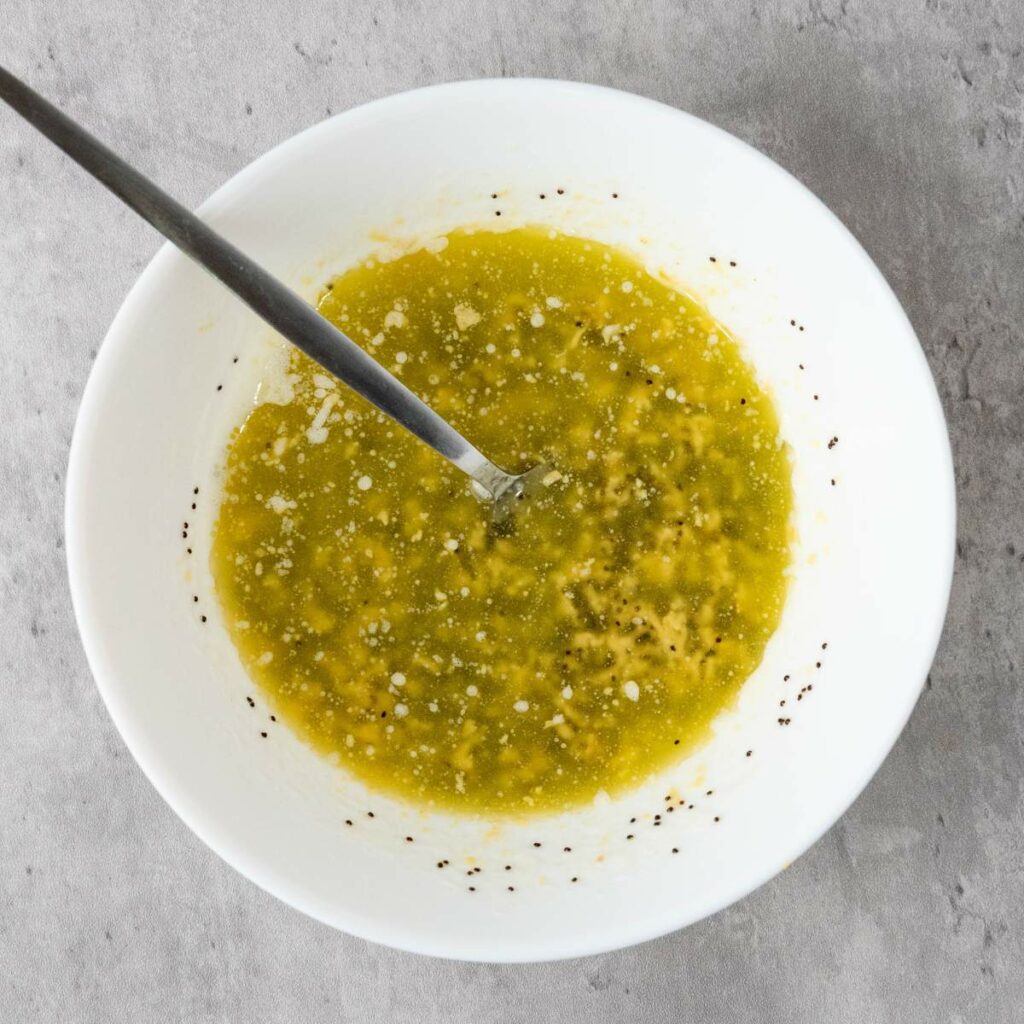 A white bowl filled with green sauce, possibly salsa, with seasoning specks, and a metal spoon on a gray surface.