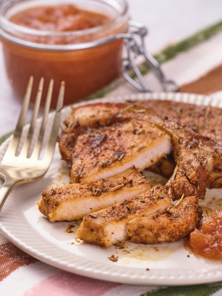 Sliced, seasoned pork chop on a white plate with a gold fork and a side of applesauce.