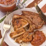 A plate with seasoned, sliced pork chop, a serving of applesauce, a fork, a knife, and a jar of applesauce in the background on a striped cloth.