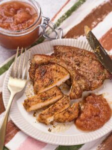A plate with seasoned, sliced pork chop, a serving of applesauce, a fork, a knife, and a jar of applesauce in the background on a striped cloth.