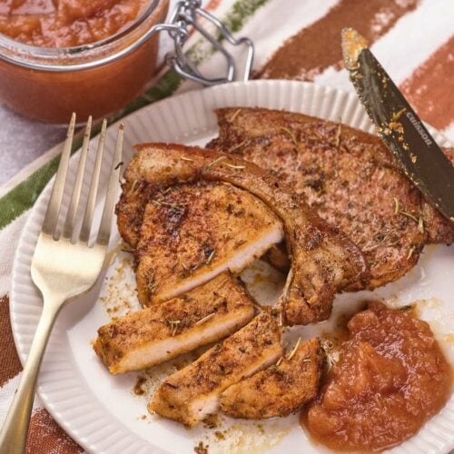A plate with seasoned, sliced pork chop, a serving of applesauce, a fork, a knife, and a jar of applesauce in the background on a striped cloth.