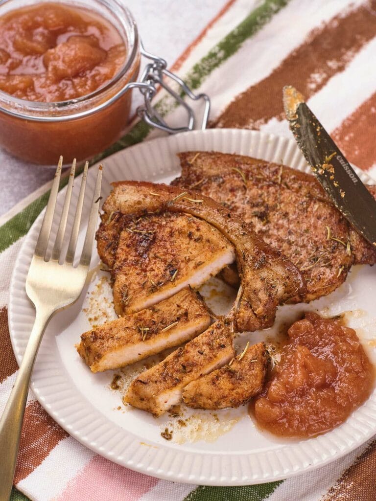 A plate with seasoned, sliced pork chop, a serving of applesauce, a fork, a knife, and a jar of applesauce in the background on a striped cloth.
