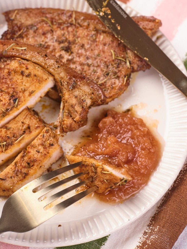 A cooked pork chop sliced on a white plate, with a fork holding a piece and a serving of applesauce on the side.