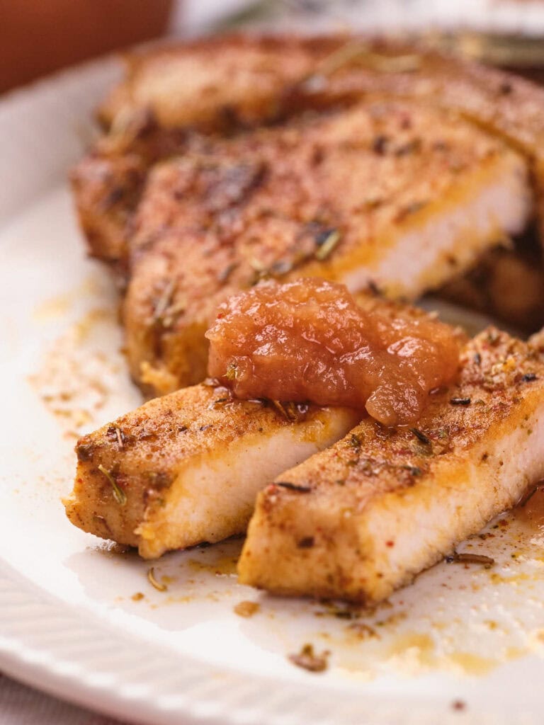 Close-up of seasoned pork chops on a plate, with two sliced pieces topped with a dollop of chunky apple sauce.