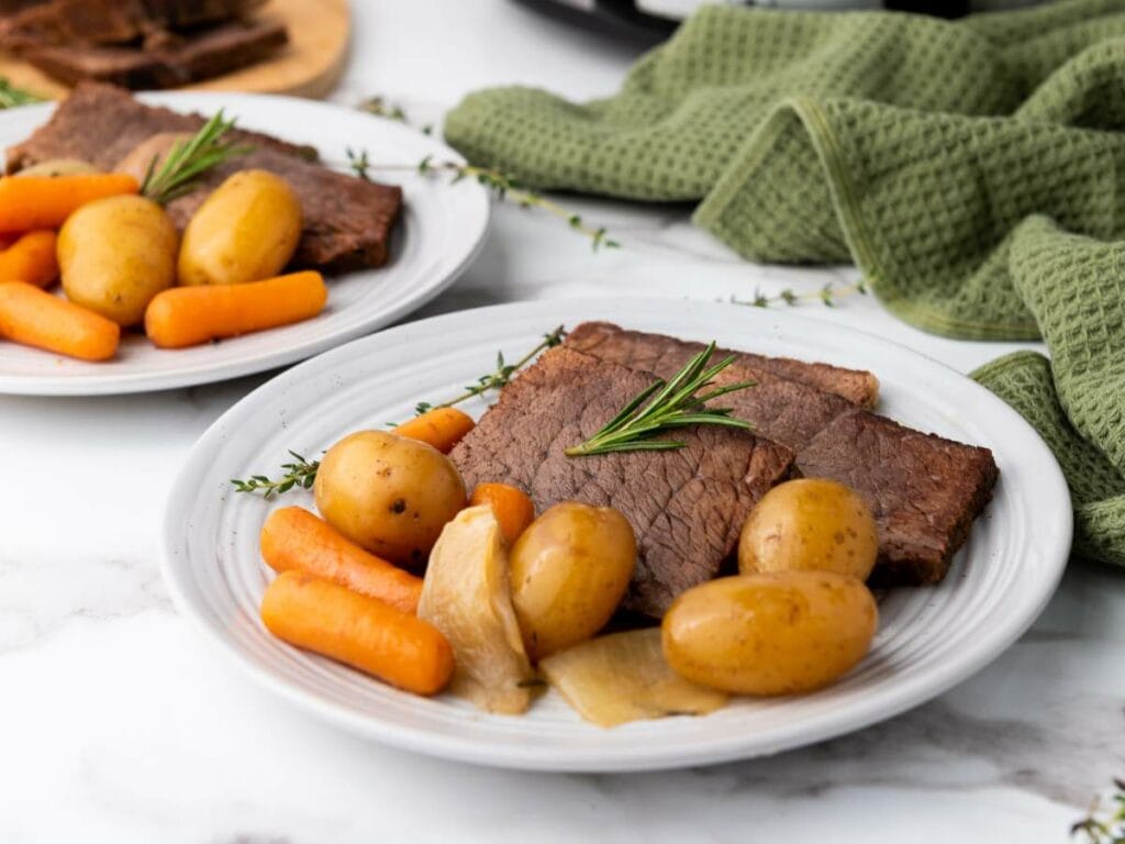 A plate with slices of rump roast, baby potatoes, carrots, and onions, garnished with rosemary, set on a white surface with a green towel in the background.
