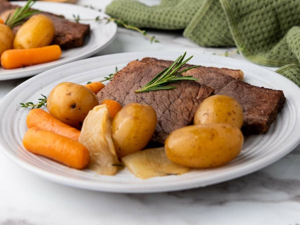 A white plate with slices of rump roast, baby potatoes, carrots, and onions, garnished with a sprig of rosemary.