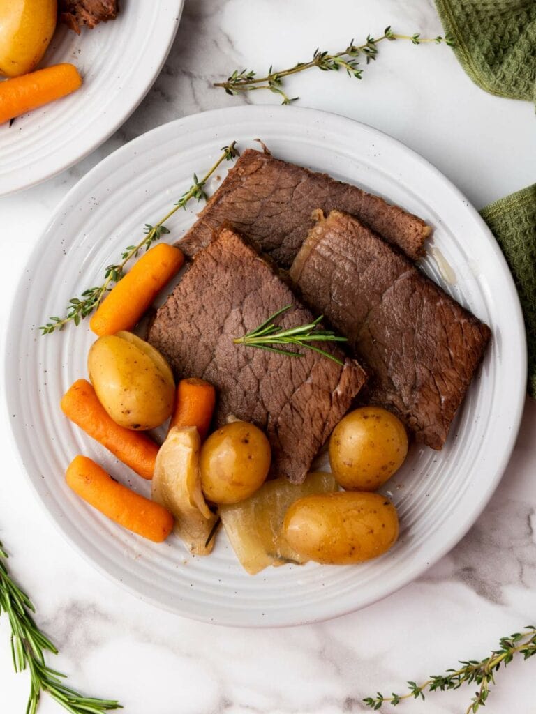 A white plate with slices of cooked rump roast, baby carrots, small potatoes, onions, and a sprig of rosemary.