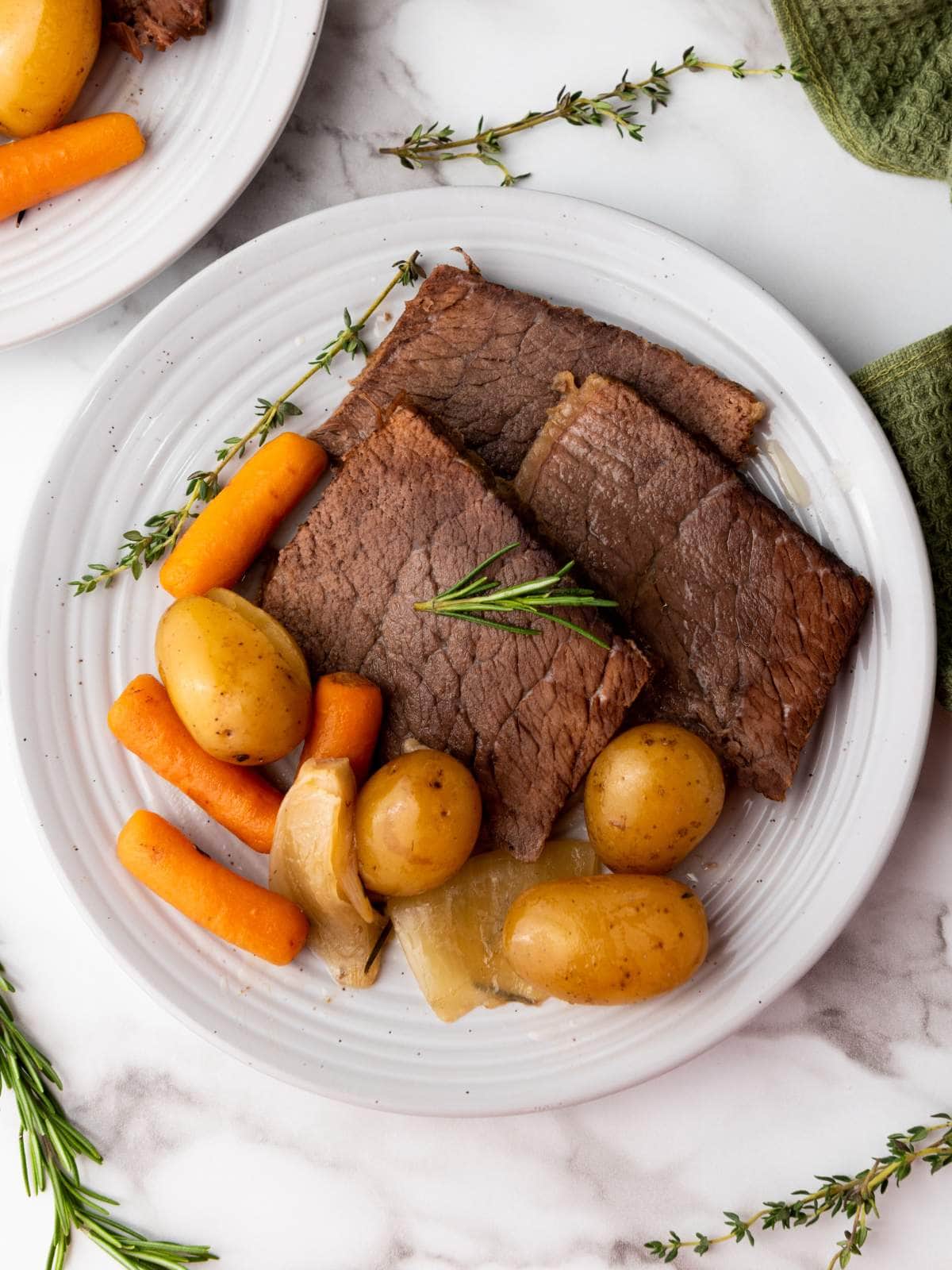 A white plate with slices of cooked rump roast, baby carrots, small potatoes, onions, and a sprig of rosemary.