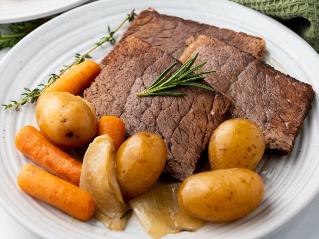 Two slices of cooked rump roast on a plate, served with baby potatoes, carrots, onion, and garnished with a sprig of rosemary.