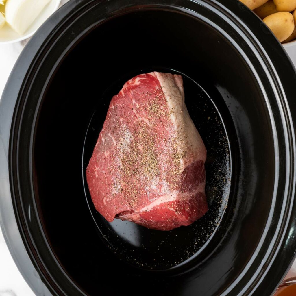 A raw seasoned beef roast sits in a black slow cooker, with sliced onions and whole potatoes nearby.