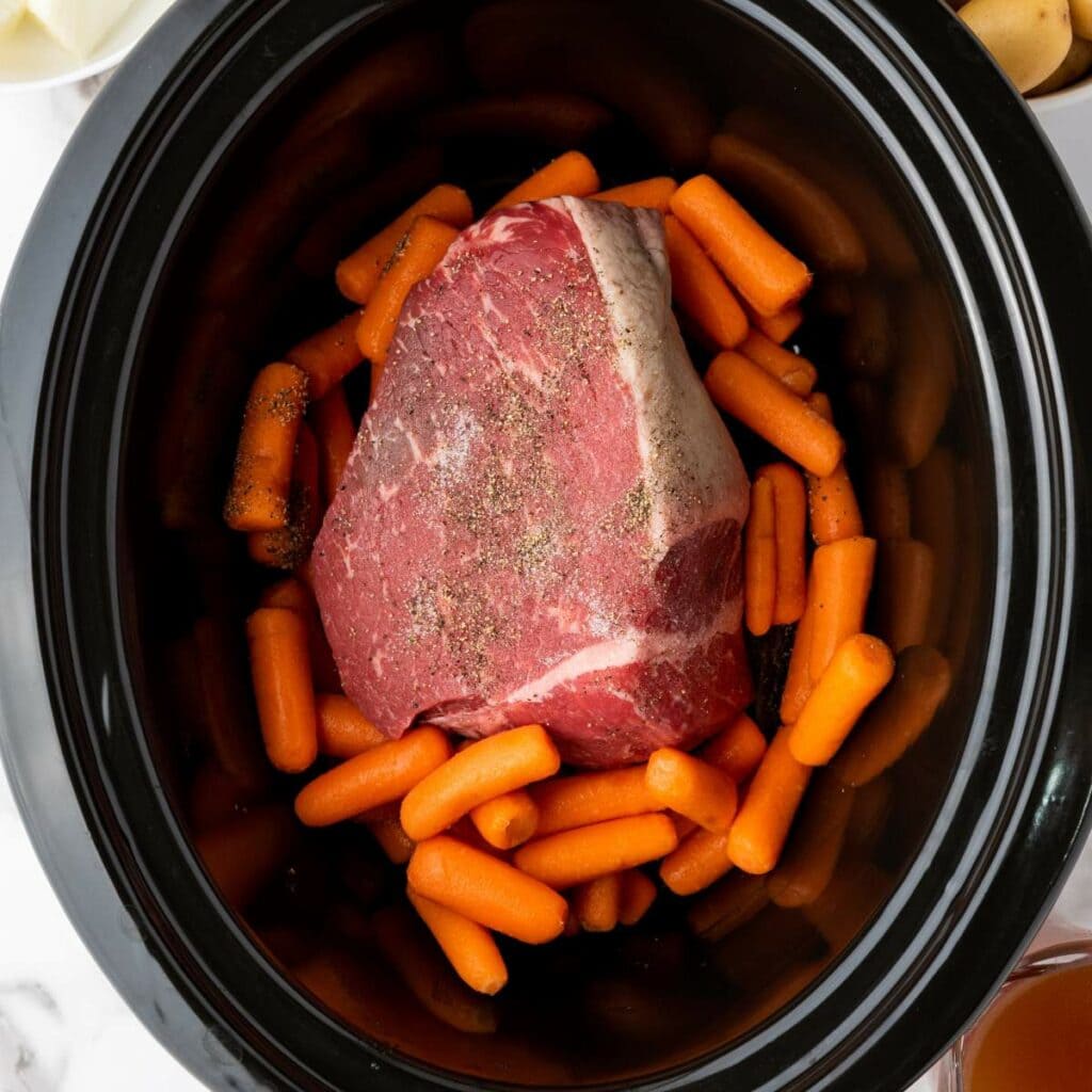 Raw beef roast seasoned with pepper sits on top of baby carrots inside a slow cooker, ready for cooking.