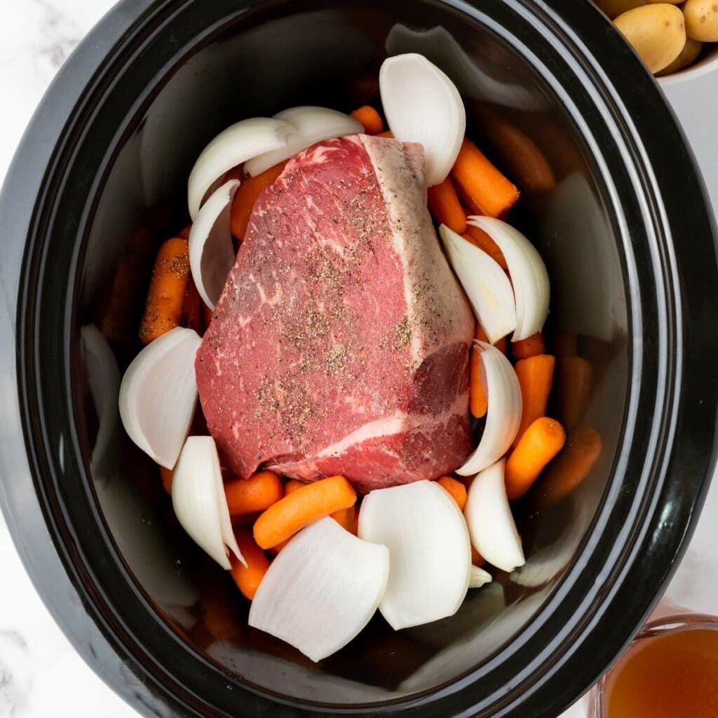 Raw beef roast seasoned with pepper, surrounded by chopped onions and baby carrots inside a black slow cooker.