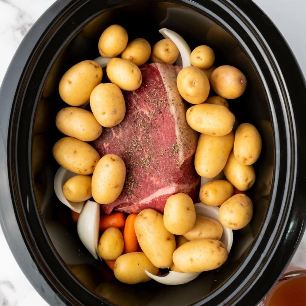 A raw beef roast, baby potatoes, carrots, and onions are arranged in a slow cooker, ready for cooking.
