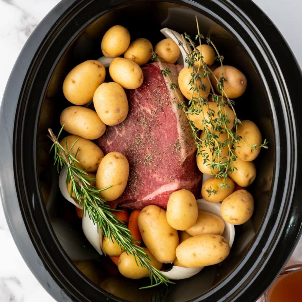 Raw beef roast, whole baby potatoes, onions, carrots, rosemary, and thyme arranged in a black slow cooker, ready for cooking.