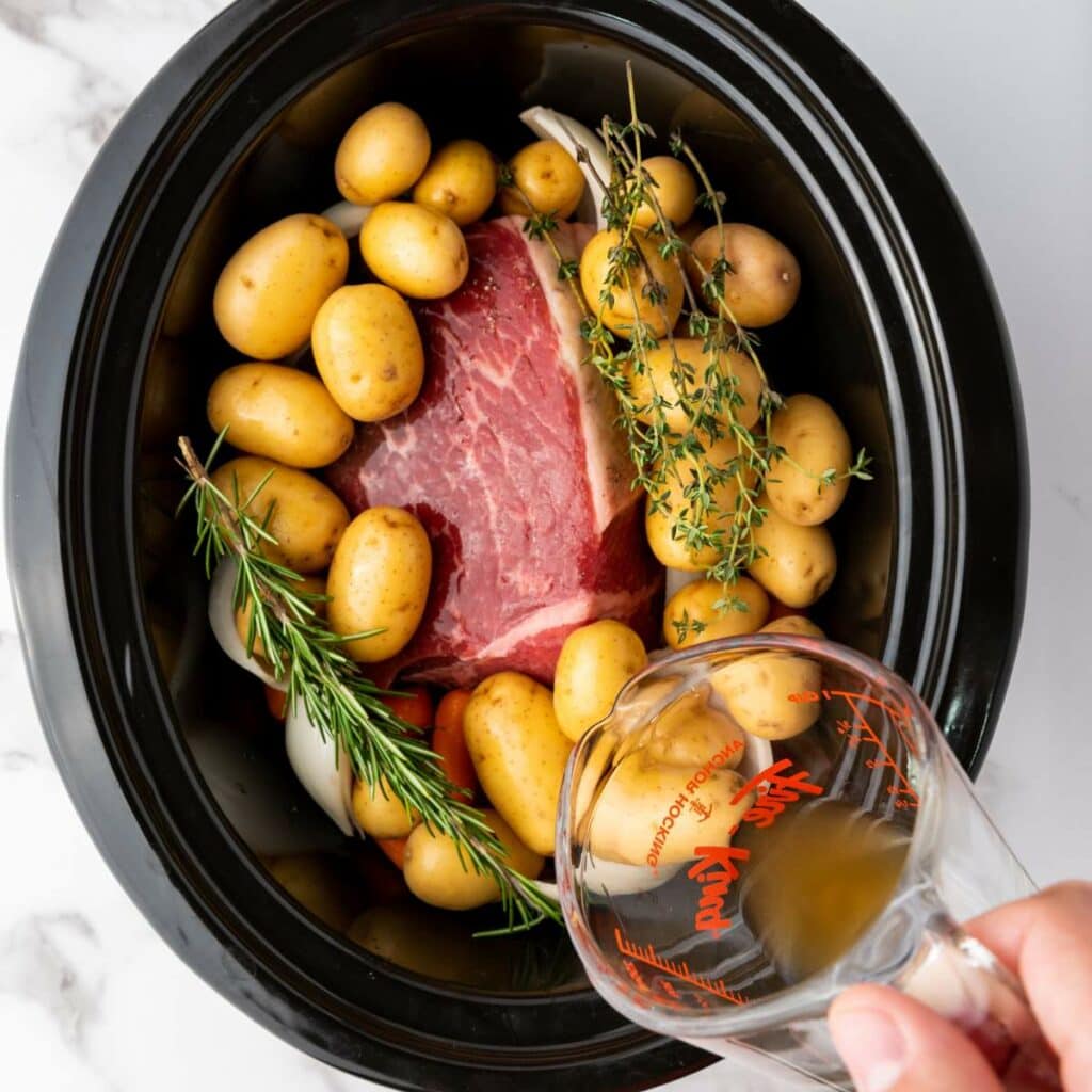 Raw beef roast, baby potatoes, herbs, and onions in a slow cooker, with broth being poured from a measuring cup over the ingredients.