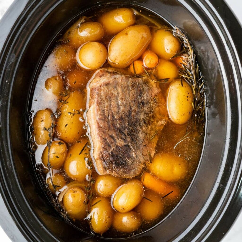 A slow cooker filled with cooked pot roast, baby potatoes, carrots, broth, and herbs.