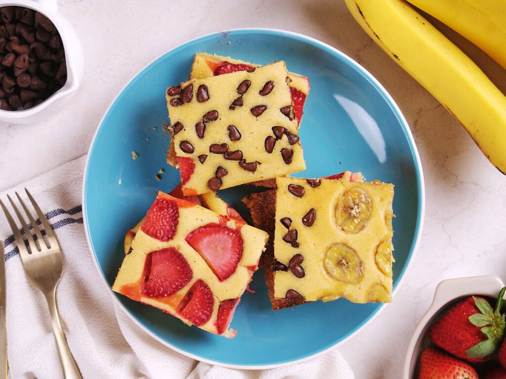 Four pieces of sheet pan pancakes topped with strawberries, chocolate chips, and banana slices on a blue plate.